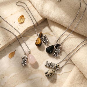 May include: Several necklaces with silver chains and gemstone pendants. The pendants feature teardrop and round stones, plus clusters of small silver beads. Gemstones are in pink, brown, black, and white. The necklaces are displayed on a cream-colored surface.