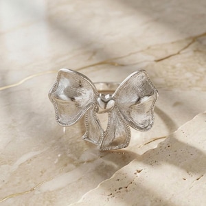 May include: A silver-toned ring featuring a prominent bow design with a textured, brushed metal finish. At the center of the bow, a small, smooth, polished heart element connects the two loops. The ring band is thin and circular, supporting the bow.