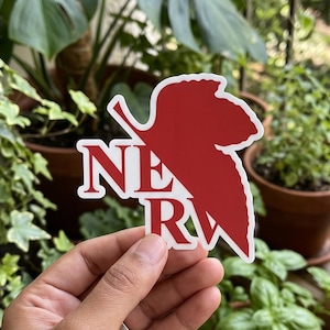 May include: A red and white sticker featuring the text "NERV" in a bold, serif font. The text is overlaid on a red leaf-shaped design. The sticker is held in a hand against a background of potted plants.
