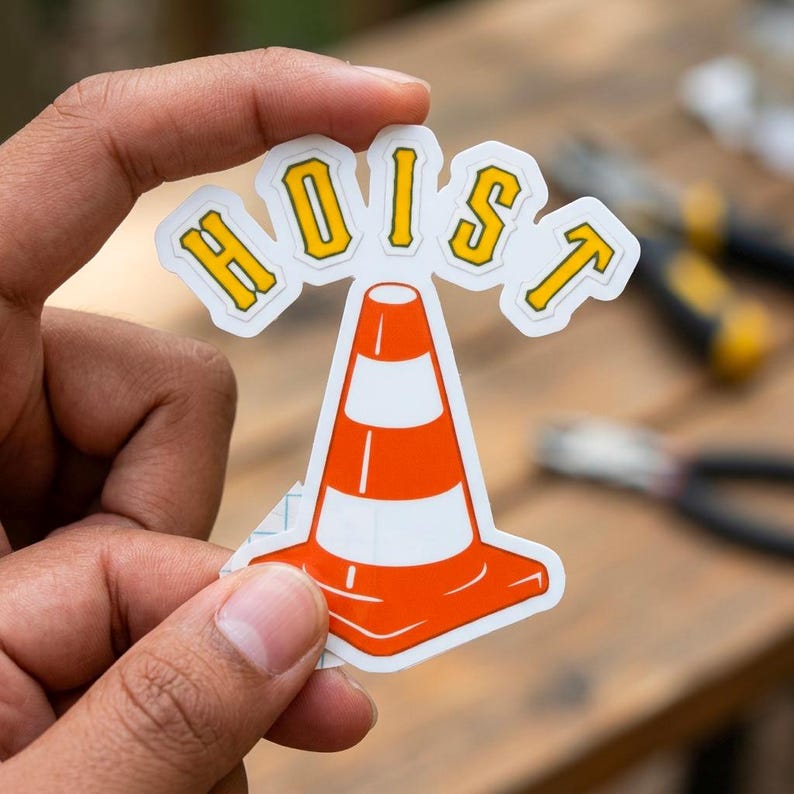 Puede incluir: Una pegatina con un dise&ntilde;o de cono de tr&aacute;fico naranja y blanco con la palabra "HOIST" en letras amarillas con un contorno blanco. La pegatina est&aacute; sujeta en la mano de una persona.