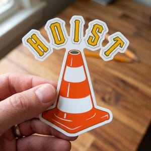 Puede incluir: Una pegatina con un dise&ntilde;o de cono de tr&aacute;fico naranja y blanco con la palabra "HOIST" en letras amarillas. La pegatina est&aacute; sujeta por una mano y el fondo es una superficie de madera.
