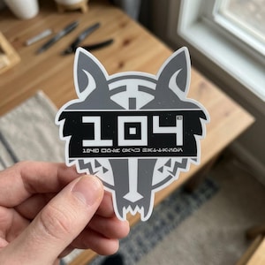 May include: A gray and white sticker featuring a wolf head design. The sticker has the number "104" in a bold, square font, with additional text below. The design is a stylized, geometric representation of a wolf.