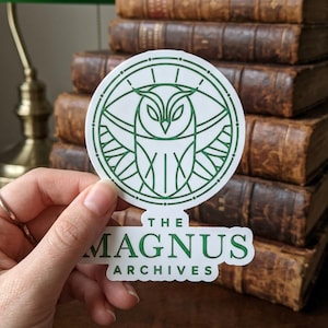 May include: A white sticker with a green owl design inside a circle, with the text "THE MAGNUS ARCHIVES" below. The sticker is held in front of a stack of old books.