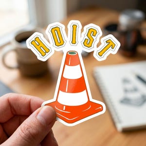 Puede incluir: Una pegatina con un cono de tr&aacute;fico naranja y blanco con la palabra "HOIST" en letras amarillas. La pegatina est&aacute; sujeta por una mano, con un fondo borroso de una taza y un cuaderno.