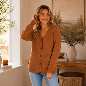 May include: A long-sleeved, rust-colored knit cardigan featuring a V-neckline and a front button closure with five dark buttons. The garment has a textured, chunky knit pattern, ribbed cuffs, and a ribbed hemline, paired with light blue denim jeans.