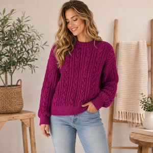 Pode incluir: Uma camisola de manga comprida em magenta vibrante com um padrão de malha de tranças vertical proeminente, gola redonda clássica e punhos e bainha canelados, combinada com calças de ganga azul-claras. A peça tem um corte descontraído.