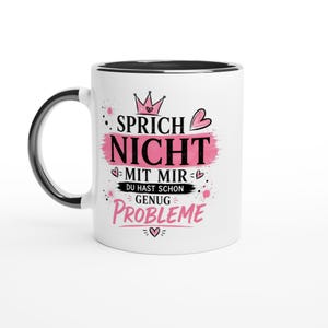 Può includere: Una tazza in ceramica bianca con manico e bordo interno neri. La parte anteriore presenta un'icona a forma di corona rosa, cuori rosa e il testo nero: SPRICH NICHT MIT MIR DU HAST SCHON GENUG PROBLEME, accentuato da pennellate rosa e linee.