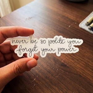 May include: A white die-cut sticker featuring black cursive lettering that reads, "never be so polite you forget your power." The sticker has a smooth, irregular outline that follows the shape of the text, creating a clean and legible design.
