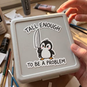 May include: A square light gray plastic container featuring a sticker of a cartoon penguin holding a large kitchen knife. The text above the penguin reads TALL ENOUGH and the text below reads TO BE A PROBLEM in bold black capital letters.