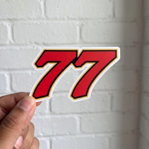 May include: A die-cut sticker featuring the number 77 in a bold, slanted font. The numerals are bright red with a thick black outline and a thin yellow border, creating a high-contrast graphic design suitable for application on various surfaces.