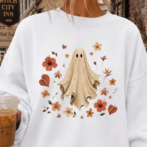 Könnte beinhalten: Ein weißes Langarm-Sweatshirt mit Rundhalsausschnitt und einem zentralen Aufdruck eines beigen Geistes, der von verschiedenen orangefarbenen, braunen und gelben herbstlichen Blumen und Blättern umgeben ist. Das Kleidungsstück hat gerippte Bündchen.