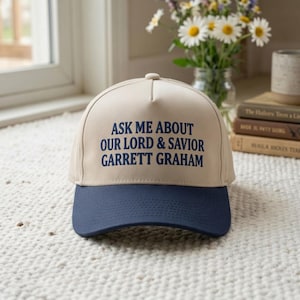 May include: A beige baseball cap with a dark blue brim and dark blue embroidered text on the front that reads: ASK ME ABOUT OUR LORD & SAVIOR GARRETT GRAHAM. The cap features a structured crown and a curved visor, providing a classic look.