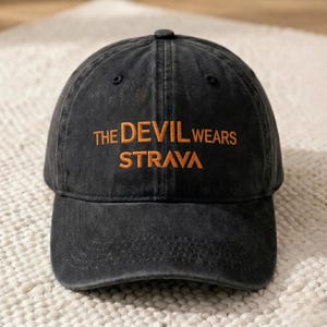 May include: A black baseball cap with the text "THE DEVIL WEARS STRAVA" embroidered in orange. The cap has a curved brim and a classic six-panel design. The hat is made of a durable, slightly faded fabric.