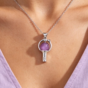 Peut inclure: Un collier pendentif argenté avec un design en forme de clé. Le pendentif a un cadre circulaire avec une sphère violette au centre. Le collier est porté autour du cou, avec la chaîne visible.