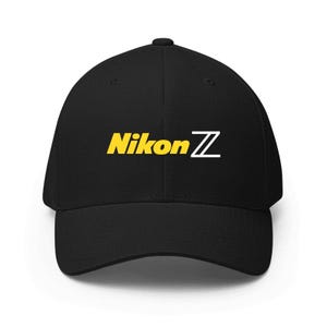 May include: A black baseball cap featuring the text Nikon Z embroidered on the front. The word Nikon is in yellow, and the letter Z is in white. The cap has a structured crown, a curved brim, and a button at the top centre of the headpiece.