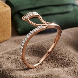 Könnte beinhalten: Roségoldener Ring mit einzigartigem Design, besetzt mit kleinen, funkelnden Steinen. Der Ring wird auf einer strukturierten, braunen Oberfläche präsentiert, mit einer braunen Schmuckschatulle und einem grünen Beutel im Hintergrund.
