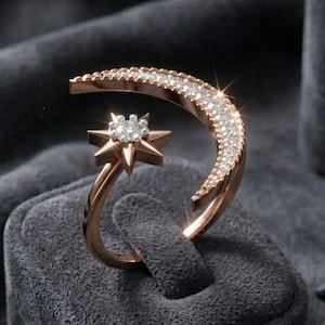Könnte beinhalten: Roségoldener Ring mit einem Halbmond-Design, besetzt mit kleinen, funkelnden Steinen. Ein sternförmiger Akzent befindet sich unter dem Mond. Der Ring wird auf einem grauen Samtständer präsentiert, was auf ein Schmuckthema hindeutet.