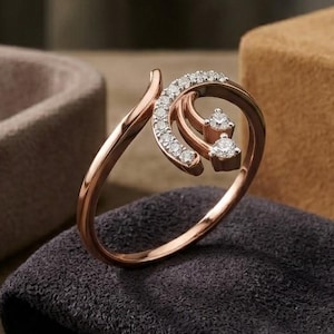 Könnte beinhalten: Roségoldener Ring mit einem wirbelnden Design, verziert mit kleinen, funkelnden Diamanten. Der Ring hat ein einzigartiges, offenes Design mit zwei größeren Diamanten, die in dem Wirbel gesetzt sind. Der Ring wird auf einem dunkelgrauen Tuch präsentiert.