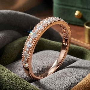 Könnte beinhalten: Roségoldener Ring mit zwei Reihen funkelnder Diamanten. Der Ring liegt auf grauem, grünem und braunem Stoff. Im Hintergrund befindet sich eine grüne Samt-Schmuckschatulle.