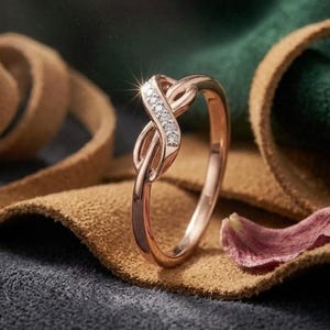 Peut inclure: Bague en or rose en forme de symbole infini, ornée d'une rangée de petits diamants blancs étincelants. L'anneau est posé sur du cuir marron, du tissu vert et une surface gris foncé.