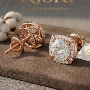 Könnte beinhalten: Ohrstecker aus Roségold mit einem quadratischen Halo aus kleinen, klaren Steinen, die einen großen, runden, klaren Stein umgeben. Die Ohrringe liegen auf einer braunen Oberfläche mit einer Baumwollpflanze und einem Etikett mit der Aufschrift "Riora".