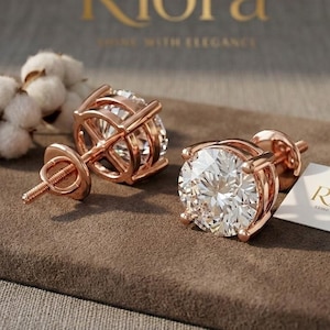 May include: Rose gold stud earrings with large, round, clear gemstones. The earrings are displayed on a brown surface with a white card that has the text "Riora" and "Shine with Elegance".