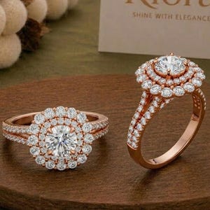 Könnte beinhalten: Zwei Ringe aus Roségold mit einem zentralen runden Stein, der von einem doppelten Heiligenschein aus kleineren runden Steinen umgeben ist. Die Ringschienen sind geteilt und mit Diamanten besetzt. Eine weiße Karte mit dem Text Riora SHINE WITH ELEGANCE ist sichtbar.