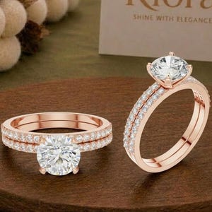 Könnte beinhalten: Ein Verlobungsring-Set aus Roségold mit einem großen runden Mittelstein und einem passenden Ring, beide besetzt mit kleinen Pavé-Diamanten. Die Ringe werden zusammen präsentiert und zeigen das roségoldene Metallfinish und klare Steine.