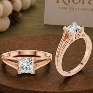 May include: Two rose gold rings featuring a square princess-cut centre stone held by a four-prong setting. The band has a split shank design that tapers towards the stone. A white card in the background displays the text Riora SHINE WITH ELEGANCE.