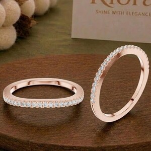 Könnte beinhalten: Zwei Ringe aus Roségold mit einem dünnen Band, das mit einer Reihe kleiner, klarer, funkelnder Steine besetzt ist. Ein Ring liegt flach auf einer runden Holzoberfläche, während der andere daneben aufrecht steht. Eine Karte mit dem Text Riora SHINE WITH ELEGANCE ist sichtbar.