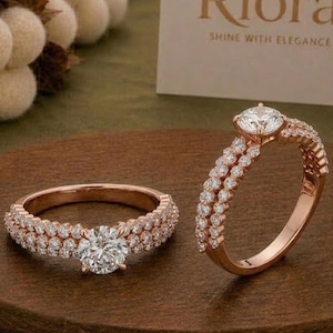 Könnte beinhalten: Zwei Ringe aus Roségold mit einem zentralen, rund geschliffenen weißen Stein und doppelten Ringschienen, die mit kleinen klaren Steinen besetzt sind. Eine weiße Karte im Hintergrund zeigt den Text Riora SHINE WITH ELEGANCE in braun.