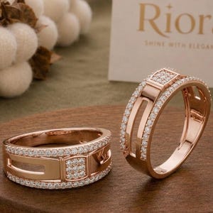 May include: Two rose gold rings featuring a wide band design with a central square section encrusted with small clear stones. The edges of the bands are lined with rows of tiny sparkling stones, creating a layered look with open cutouts.