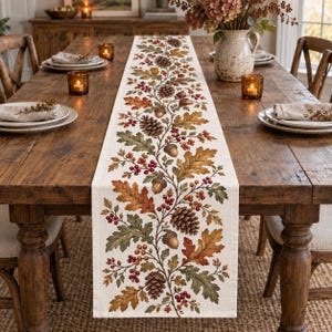 May include: A rectangular table runner featuring a printed pattern of autumn oak leaves, pinecones, and red berries on a cream-colored fabric. The botanical design runs vertically along the center, showcasing shades of green, brown, and orange.