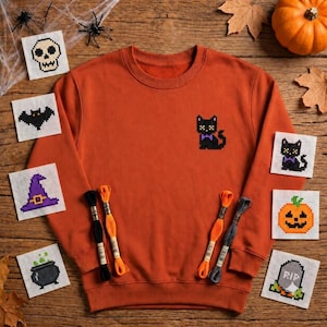 May include: A burnt orange crewneck sweatshirt featuring a small embroidered black cat with a purple bow tie on the chest. The sweatshirt is surrounded by several small square fabric patches displaying cross-stitch designs of Halloween icons.