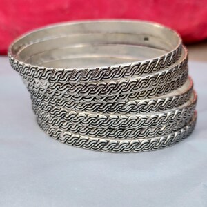 以下が含まれることがあります： 波模様が繰り返される銀色のバングルが積み重ねられています。光沢のある金属製のバングルは、テクスチャード加工のデザインです。バングルは積み重ねられており、一番上のバングルはわずかに傾いています。直径約6.5cm。