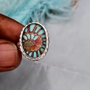 Puede incluir: Anillo de plata ovalado con un amonita fosilizada. La pieza central del anillo muestra un patrón en espiral en tonos marrón, turquesa y naranja, sobre un fondo azul claro. El anillo tiene un borde texturizado y festoneado.