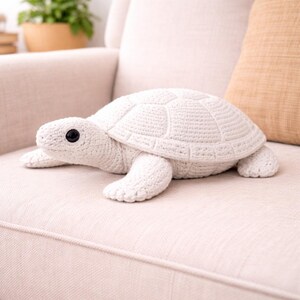 May include: A white crocheted turtle plush toy with a textured shell and black eyes. The turtle is lying on a light-colored sofa. The turtle's shell is approximately 12 inches long.