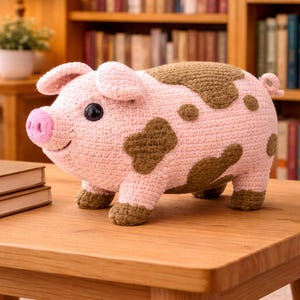 Puede incluir: Figura de cerdo de ganchillo hecha a mano en rosa y marrón. El cerdo tiene ojos oscuros, un hocico rosa y manchas marrones. Está sobre una mesa de madera. El cerdo es un objeto decorativo.