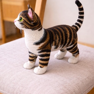 Könnte beinhalten: Gehäkelte Katzenfigur mit braunen, schwarzen und weißen Streifen. Die Katze hat grüne Augen, rosa Ohren und einen gestreiften Schwanz. Sie steht auf einem hellen Stuhlkissen. Die Katze ist handgefertigt.