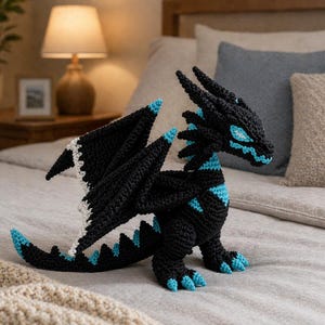 May include: A black crochet dragon with blue accents on its wings, claws, and facial features. The dragon has white trim on the wings and is posed on a bed. The dragon is a handmade item.