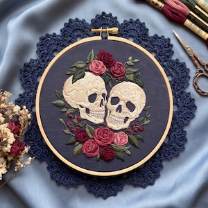 Könnte beinhalten: Ein Stickrahmen mit dunkelblauem Stoff und einem handgestickten Motiv aus zwei cremefarbenen menschlichen Schädeln, umgeben von roten und rosafarbenen Rosen mit grünen Blättern. Der Rahmen liegt auf einem blauen Satinstoff mit Stickgarn.