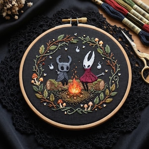 Puede incluir: Un bastidor de bordado circular de madera con tela negra que muestra una escena bordada a mano de dos personajes sentados en un tronco junto a una fogata. El diseño está rodeado por una corona de hojas verdes, ramas y setas pequeñas.