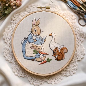 Op de afbeelding: Een ronde houten borduurring met een stoffen paneel met een gedetailleerde borduurillustratie van een konijn dat een boek voorleest aan een kleiner konijn, een witte gans en een bruine eekhoorn. De scène bevat oranje wortels en groene bladeren.