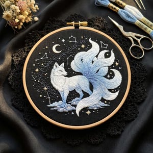 Puede incluir: Un bastidor de bordado circular de madera que contiene un panel de tela negra con un zorro de nueve colas bordado a mano en tonos blancos y azules. El zorro se encuentra sobre nubes bajo una luna creciente, estrellas y constelaciones.