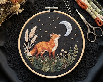 May include: An embroidery hoop containing a black fabric panel with a hand-stitched scene of a red fox standing in a field of green plants and white flowers under a crescent moon and small yellow stars, accompanied by embroidery floss and scissors.