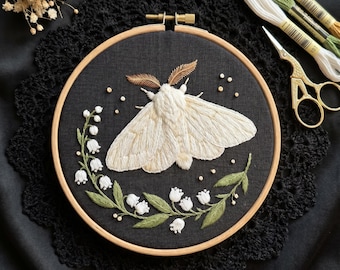 May include: A circular wooden embroidery hoop containing a black fabric panel with a hand-stitched design of a large white moth centered above a curved sprig of white lily of the valley flowers with green leaves and small scattered white beads.