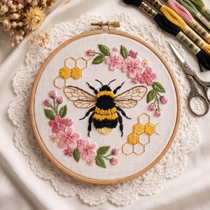 Könnte beinhalten: Ein runder Holzstickrahmen mit weißem Stoff, der ein handgesticktes Motiv einer zentralen Hummel zeigt, umgeben von rosa Blumen, grünen Blättern und gelben Wabenmustern, begleitet von Stickgarn und einer Schere.