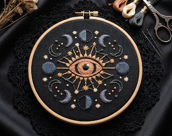 May include: A circular wooden embroidery hoop containing black fabric with a hand-stitched design of a central eye surrounded by various moon phases and small stars in shades of gold, copper, and blue thread, accompanied by embroidery floss and scissors.