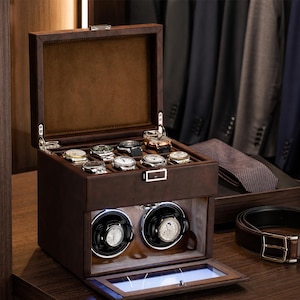 May include: A brown leather watch box with a hinged lid, holding several wristwatches. The box has a lower compartment with two watch winders. A brown belt and a tie are visible in the background.