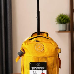 May include: A mustard yellow rolling cabin bag with a black retractable handle. The bag features a circular logo patch, side compression straps with black buckles, and a rectangular tag labelled UNDERSEAT CABIN BAG ON WHEELS with 40x25x20cm dimensions.